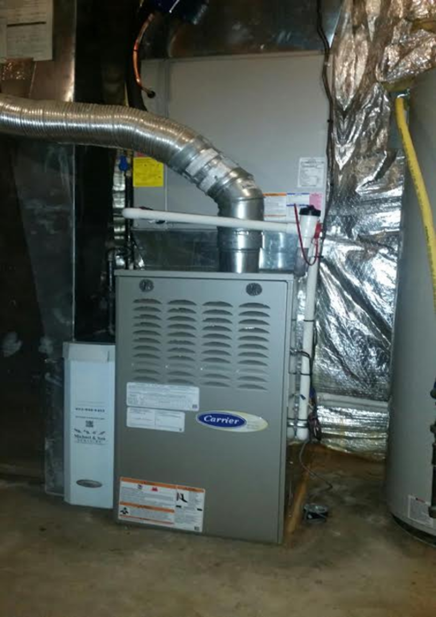 A carrier air conditioner is sitting in a basement next to a water heater.