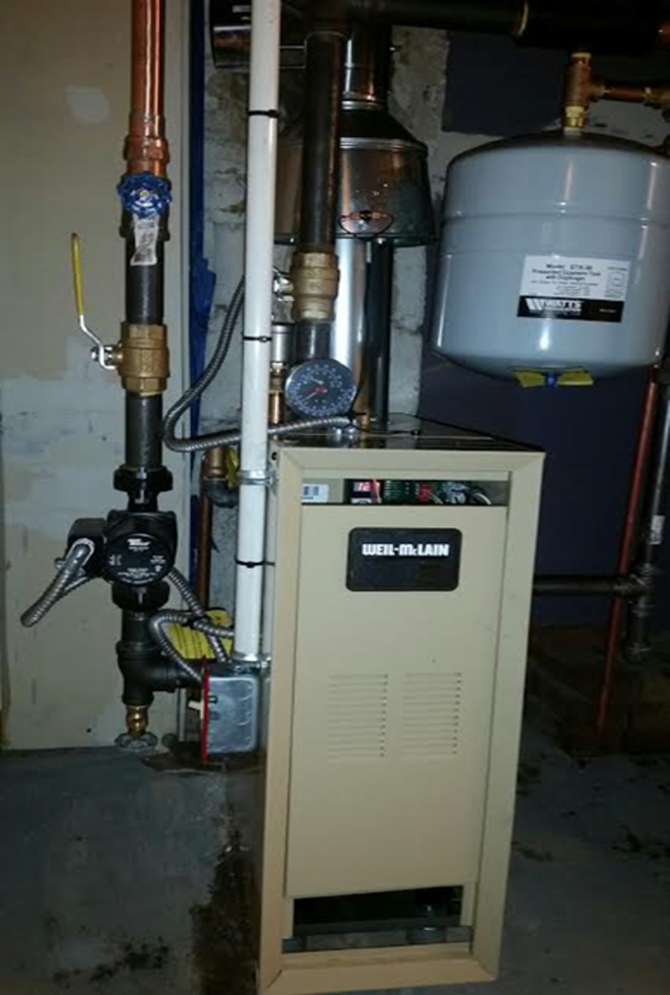 A boiler is sitting in a basement next to a water tank.