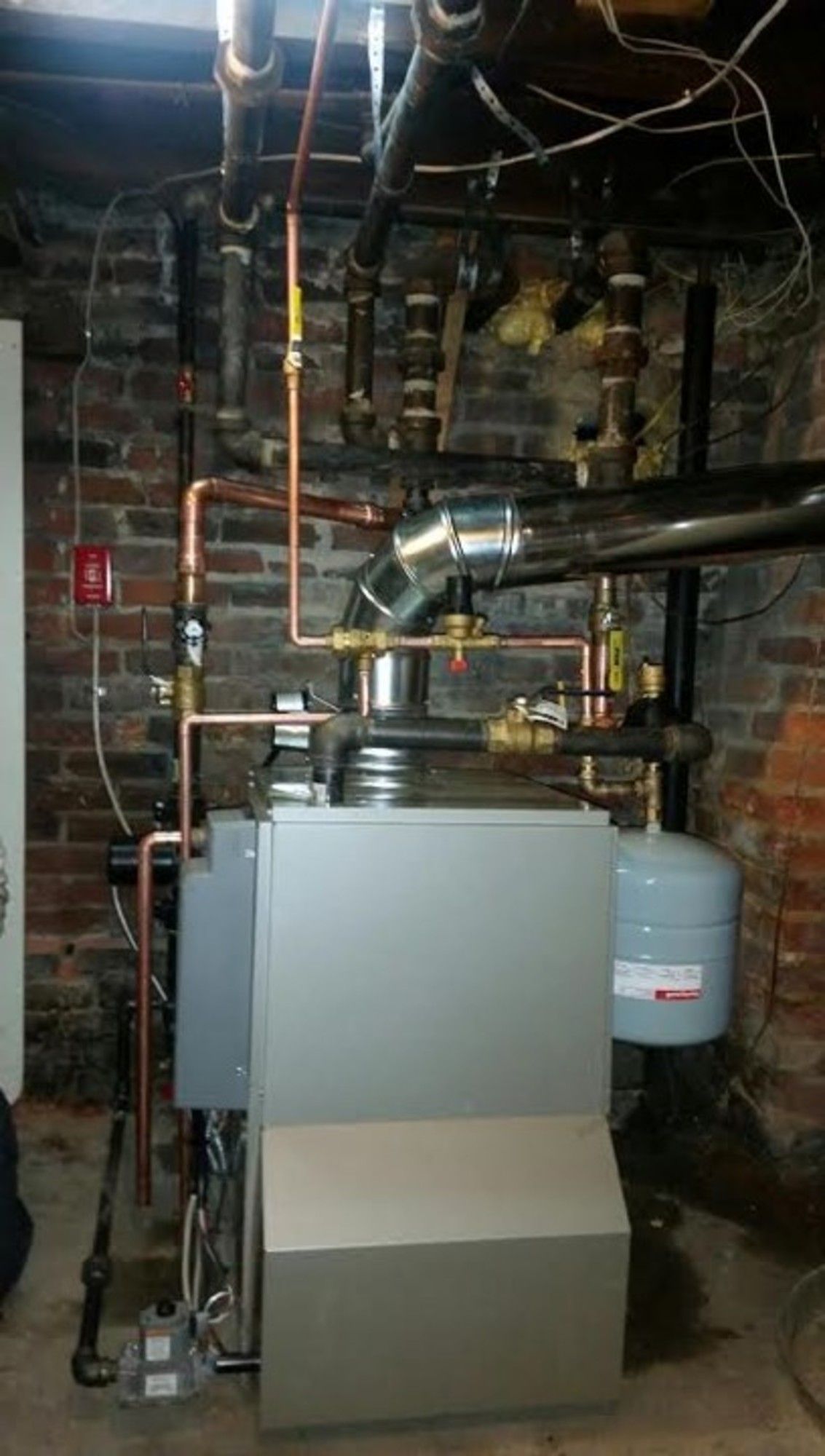 A boiler is sitting in a basement next to a brick wall.
