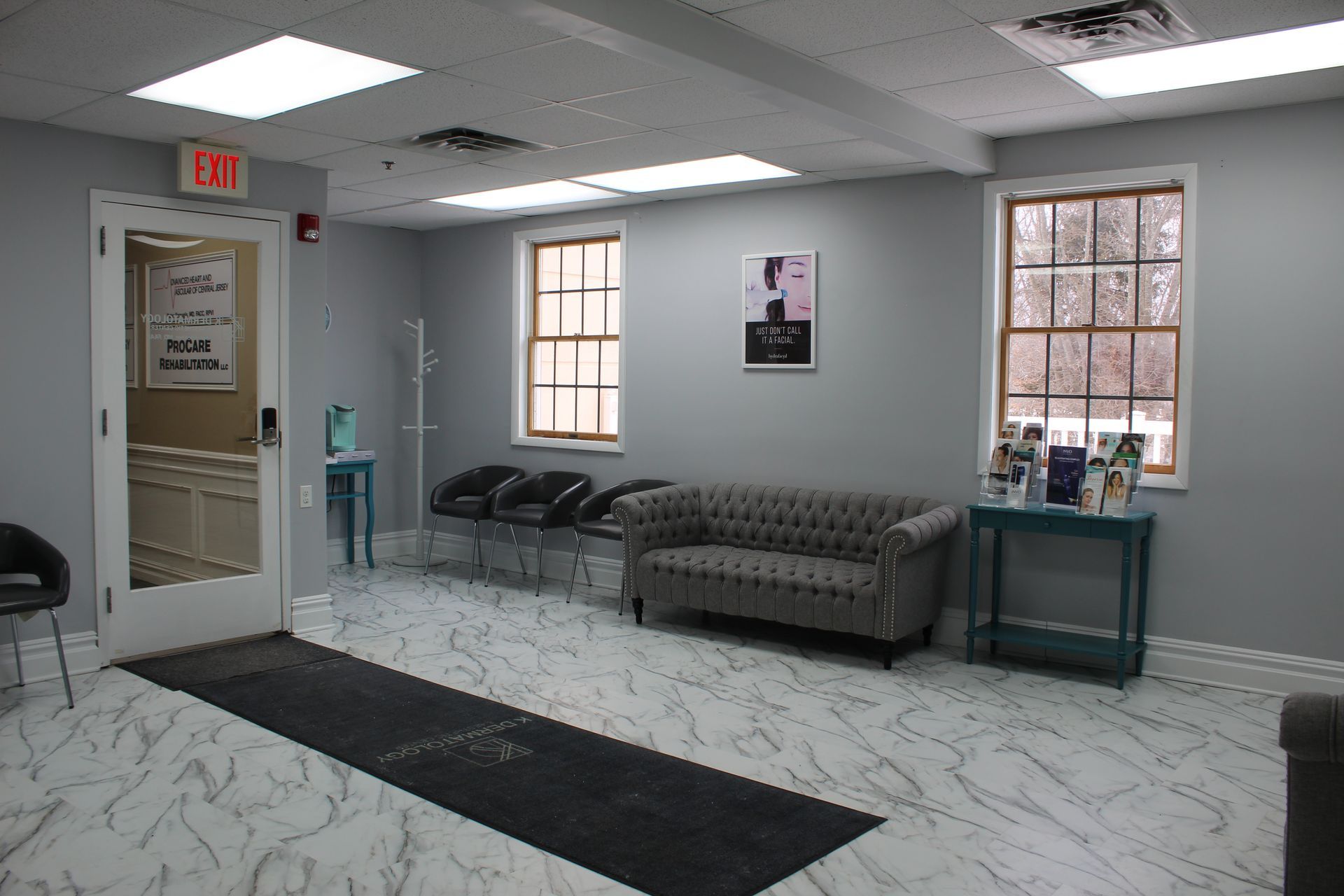 Small waiting room with gray chairs, couch, windows, and a door under fluorescent ceiling lights.