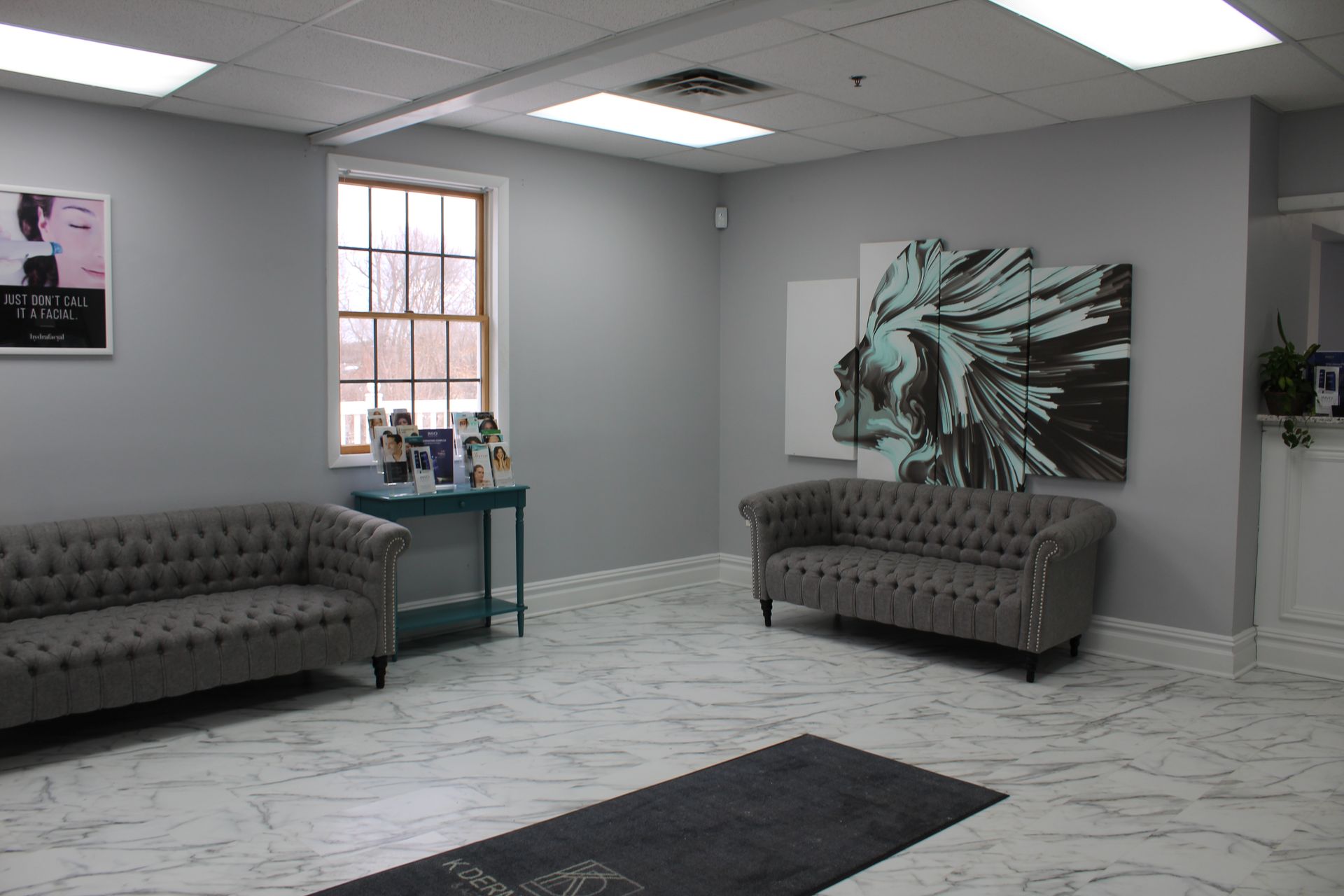 Modern waiting room with gray sofas, marble floor, wall art, and a small table by the window