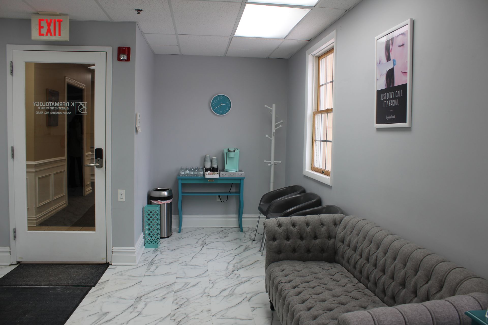 Modern clinic waiting room with tufted gray sofa, teal table, medical stand, window, and EXIT door