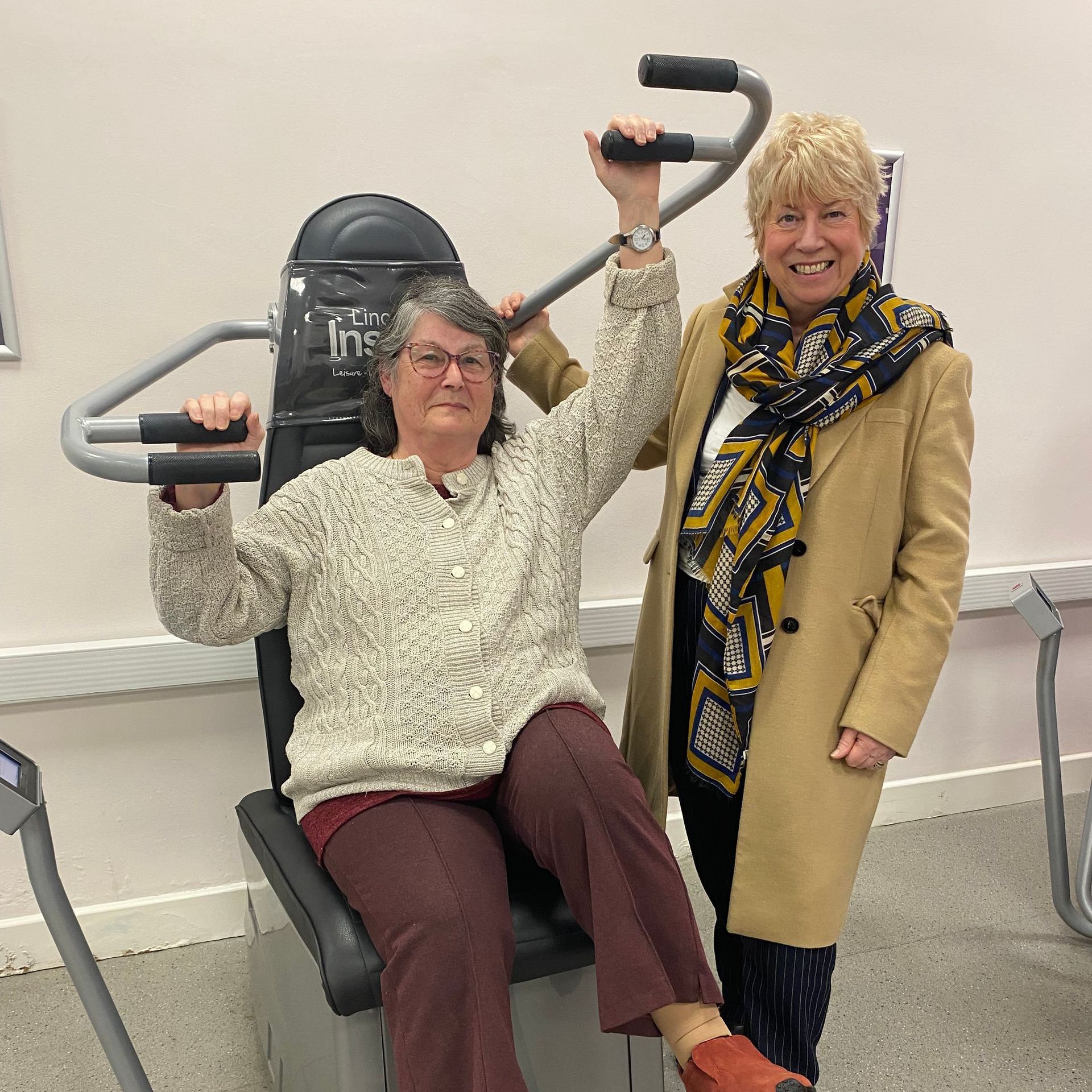 Two women are standing next to each other and one is sitting on a machine