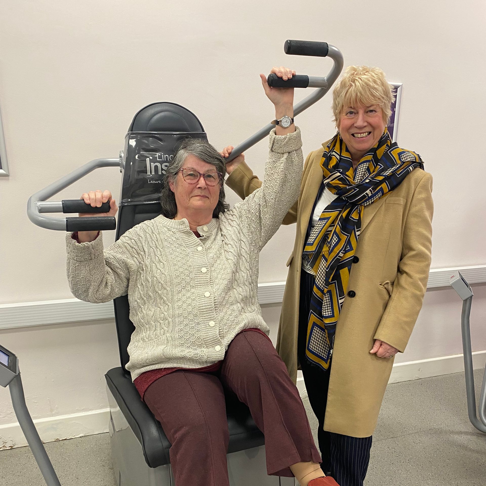 Two women are standing next to each other and one is sitting in a chair