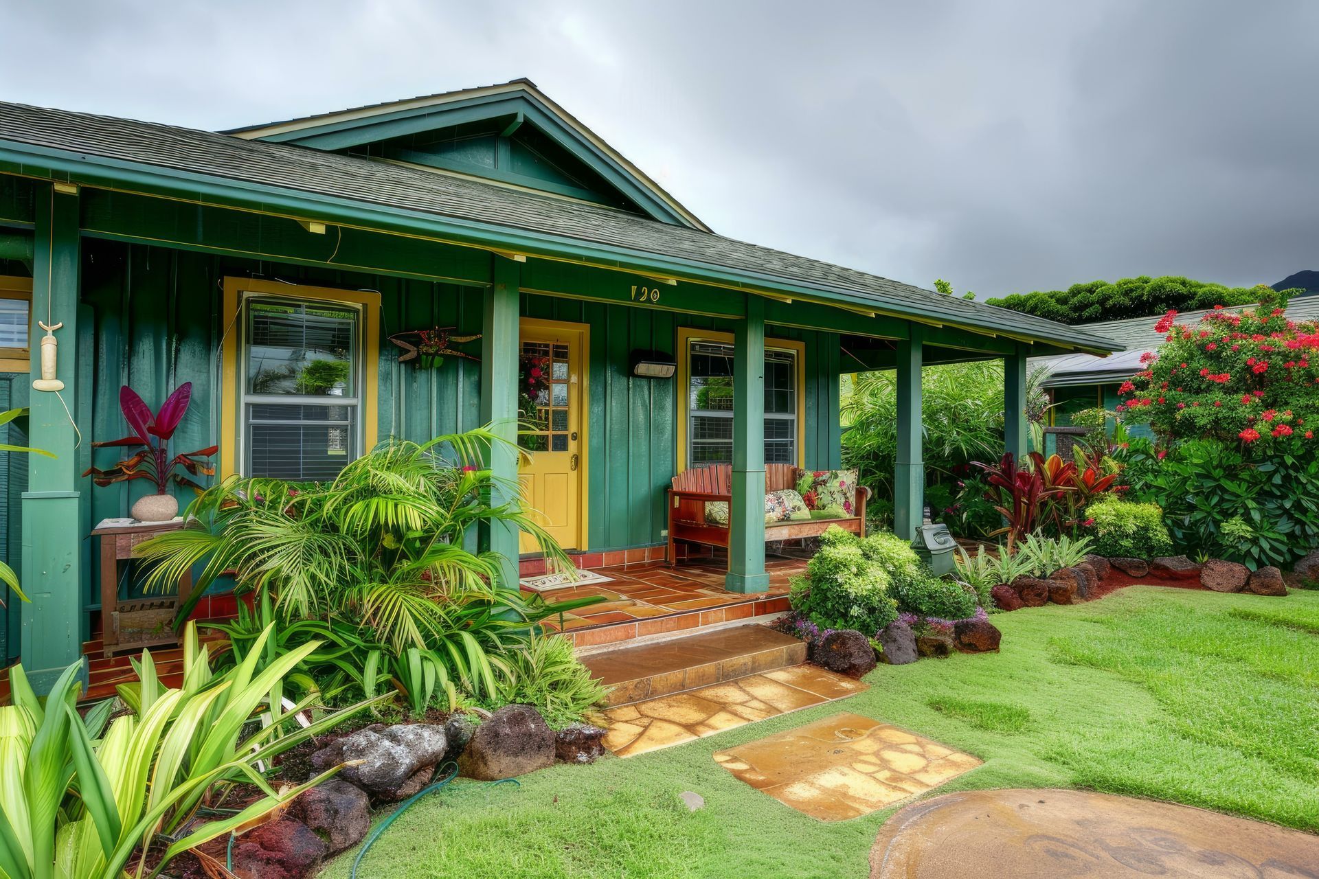 Green cottage with yellow door and porch, lush garden with red flowers and greenery.