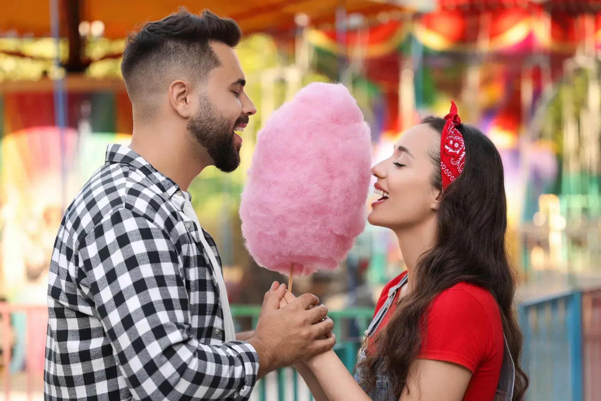 A man and a woman are eating pink cotton candy at a carnival.