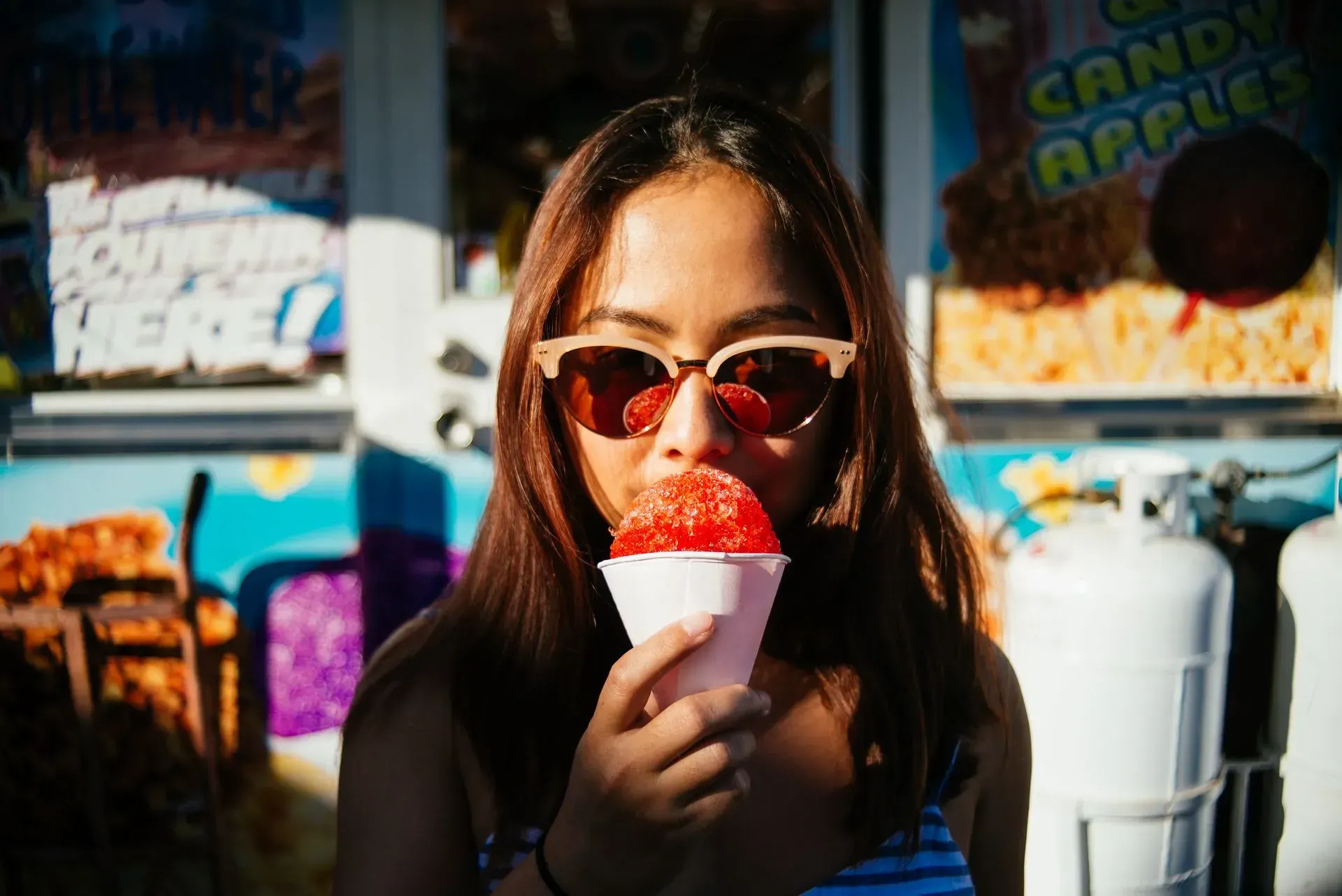 A woman wearing sunglasses is eating an ice cream cone at a carnival.