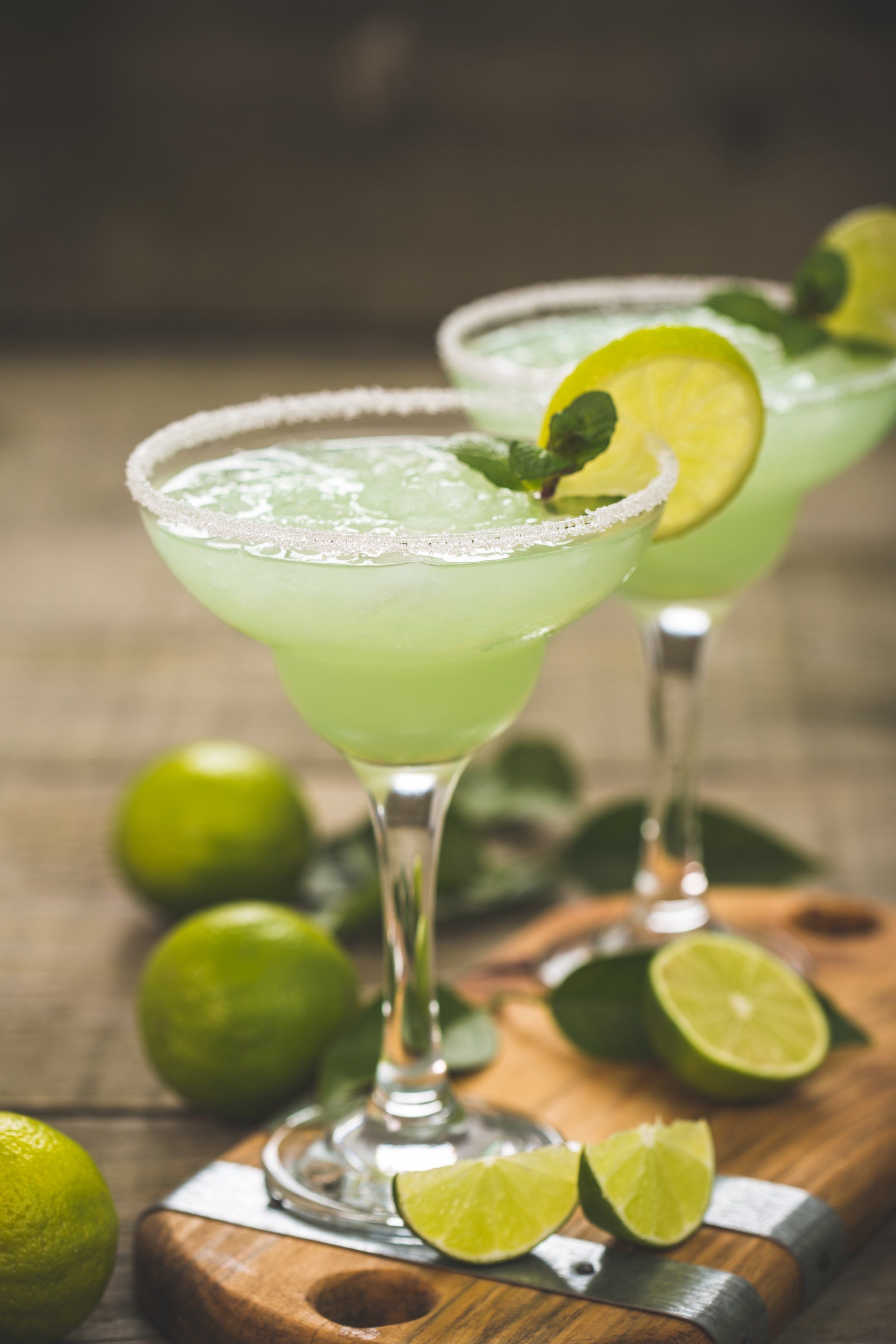 Two green margaritas with limes and mint on a wooden table.