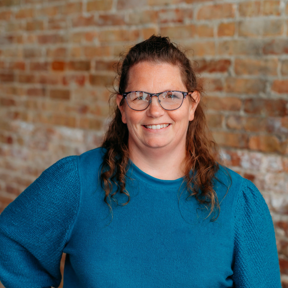 A smiling person with wavy brown hair and glasses wears a teal sweater, standing before a textured brick wall.