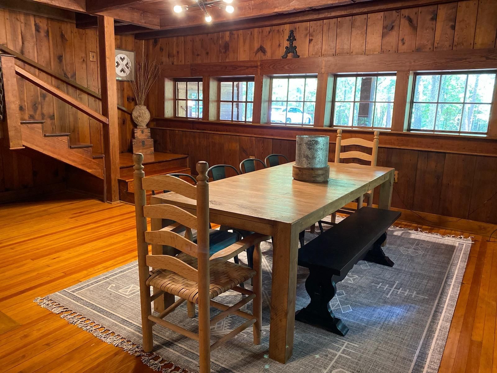 A rustic wood-paneled dining room features a wooden table with chairs and a black bench atop a grey area rug.