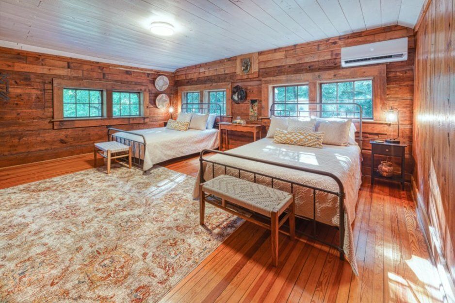 A rustic log-walled bedroom features two beds with light-colored bedding, wooden flooring, and a large patterned rug.