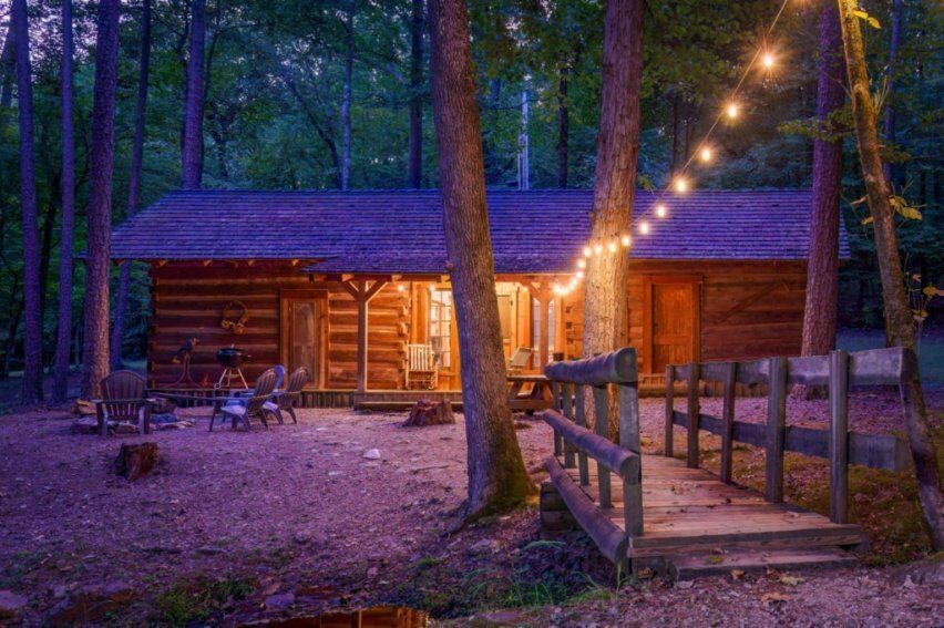 A rustic wooden cabin in a forested area at dusk, lit by warm interior lights and a string of outdoor bulbs over a bridge.