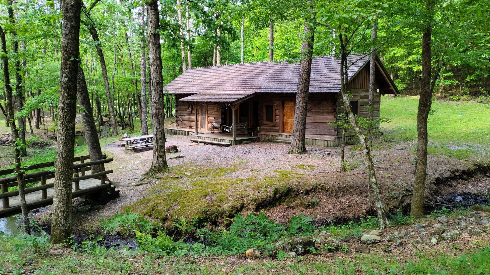 A rustic log cabin in a lush, green forest with a small wooden bridge crossing a stream in the foreground.