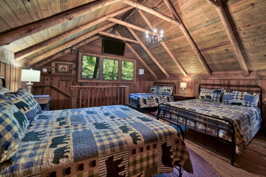 A rustic bedroom in a wooden attic featuring two beds with patterned bear quilts, exposed rafters, and a ceiling light.