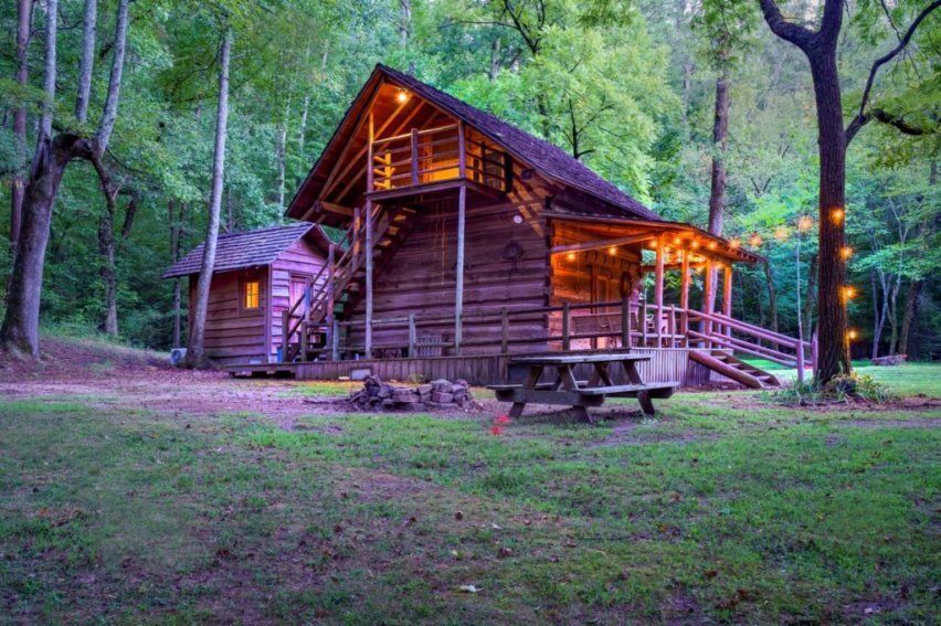 A rustic wooden cabin with a porch and lit loft stairs sits in a lush, green forest at dusk, featuring a nearby picnic table.
