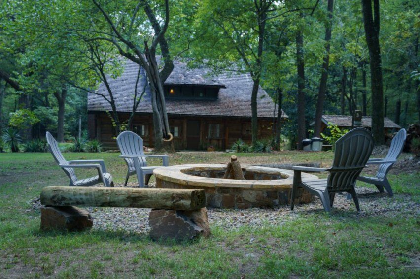 A stone fire pit surrounded by Adirondack chairs and a log bench, with a wooden cabin in a lush forest setting.