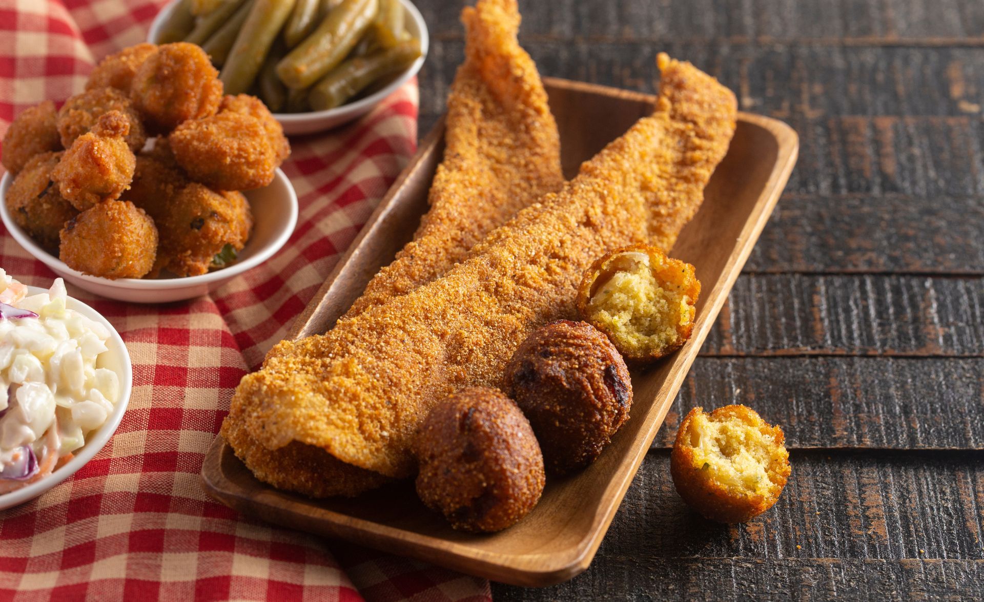 A wooden platter with breaded fried fish and hushpuppies, accompanied by bowls of coleslaw and green beans on a plaid cloth.