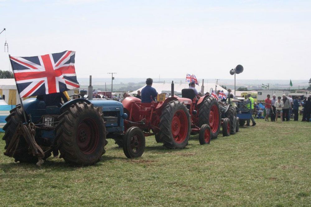 A row of tractors are parked in a field with a british flag on top of one of them.