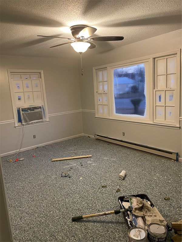 Empty room with blue carpet, gray walls, two windows, ceiling fan, and painting supplies.