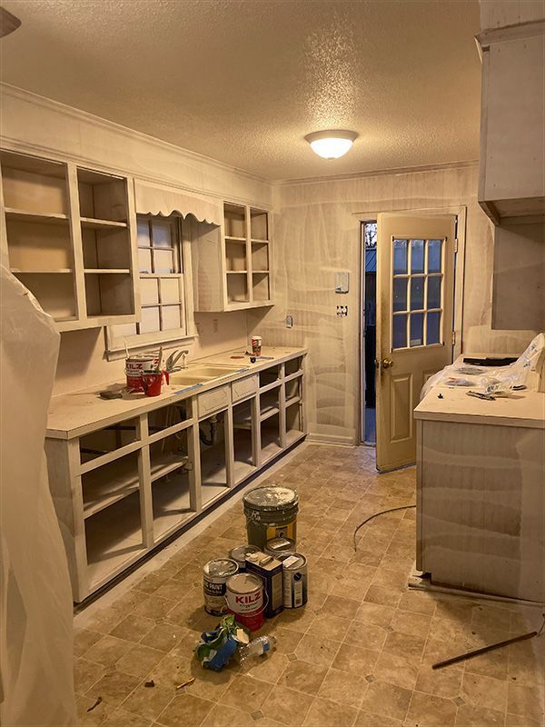 Kitchen interior during renovation with bare cabinets, paint cans, and open doorway.