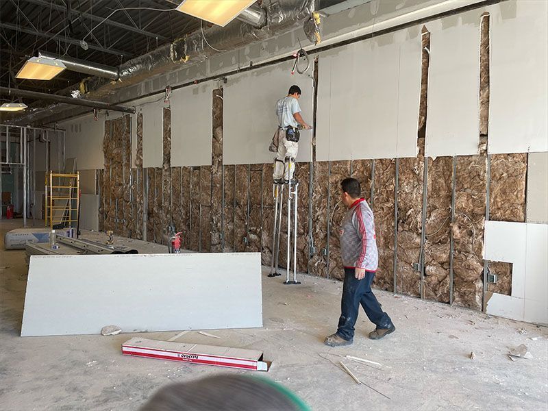 Construction workers installing drywall in a commercial space. One stands on stilts, the other walks by.