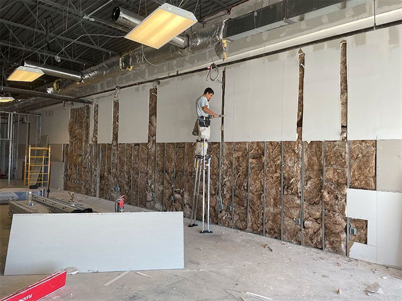 Construction worker on stilts installing drywall on a wall with insulation exposed.