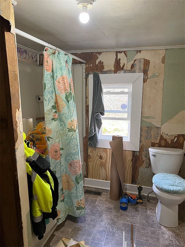 Bathroom undergoing renovation; exposed walls, floral shower curtain, toilet, window, work clothes hanging.