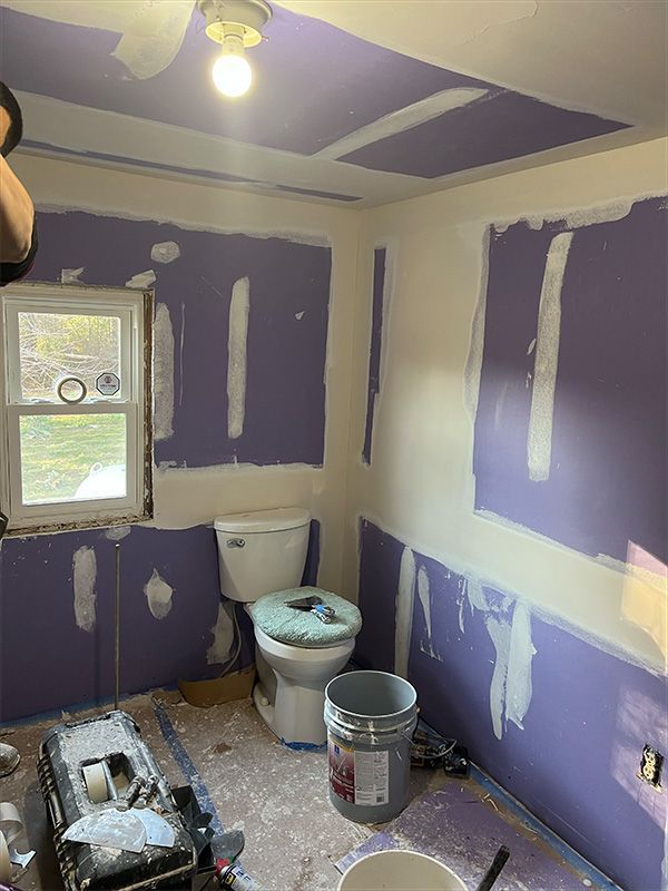 Bathroom under renovation, purple drywall with white joint compound applied, toilet, window, bucket.