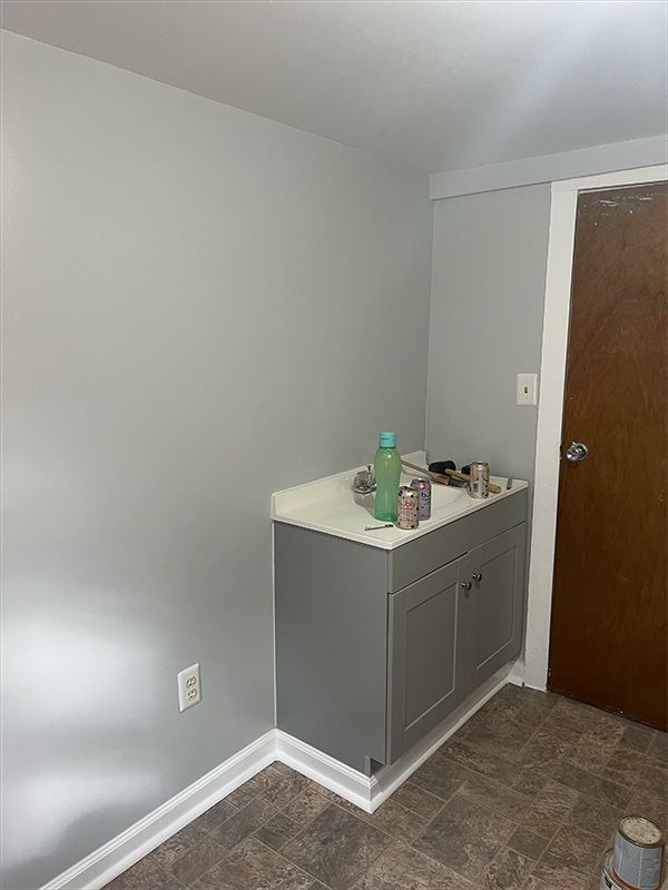 Gray cabinet with white countertop in a corner next to a closed brown door and gray walls.