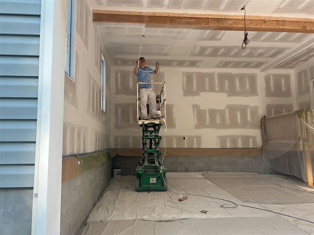 A person on a lift paints a drywall ceiling in a room with bare concrete floor and a wooden beam.