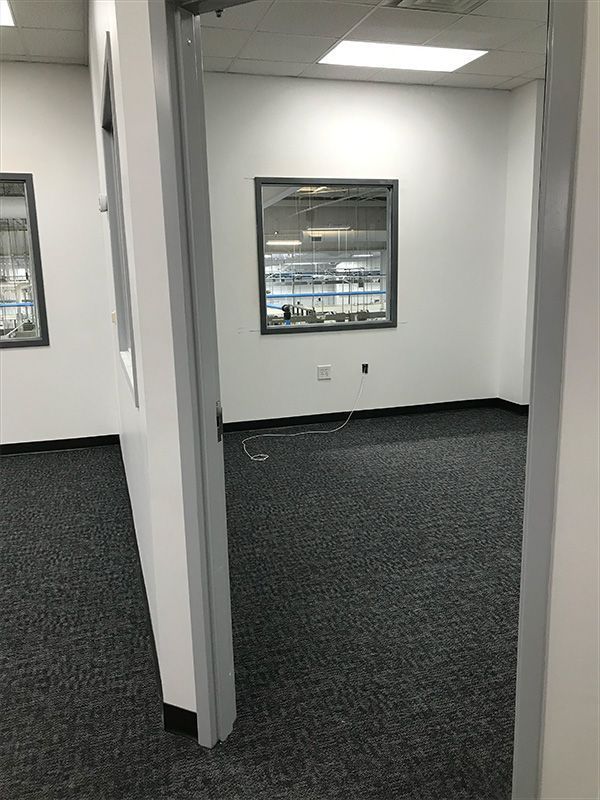 Empty office space with gray carpet, white walls, and a window looking into another room.