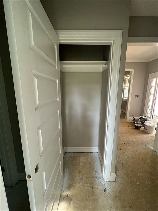 Open white closet door, closet with a hanging rod, gray walls, and unfinished floor in a doorway.