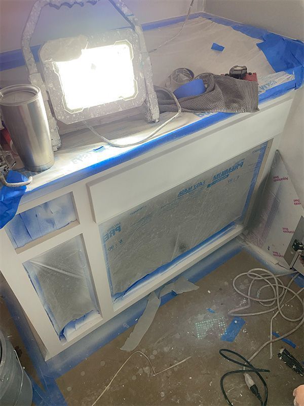 White cabinet being prepped for painting, covered in blue painter's tape, lit by a work light.