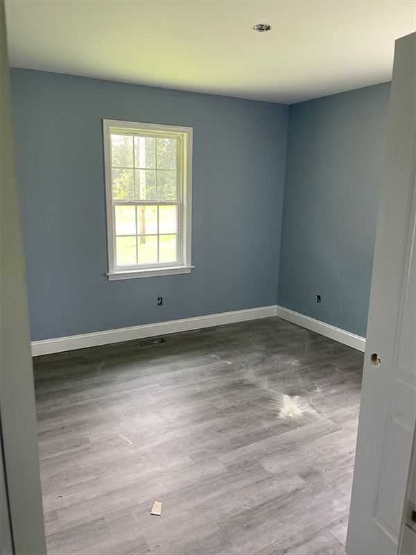 Empty bedroom with blue walls, white trim, window, and gray flooring.