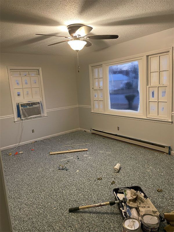 Empty room with light gray walls, blue and gray carpet, white trim, two windows covered, ceiling fan, and painting supplies.