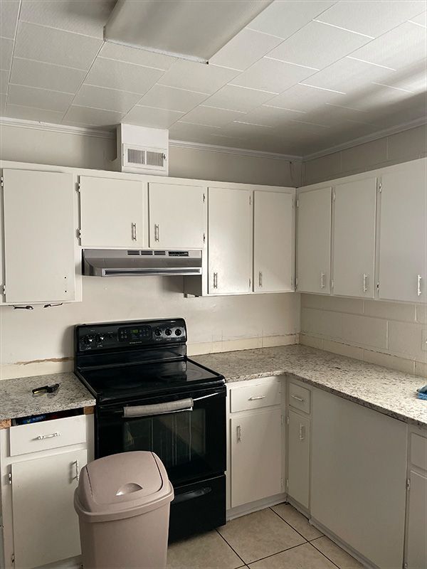 White kitchen cabinets surround a black stove and tan trash can. 