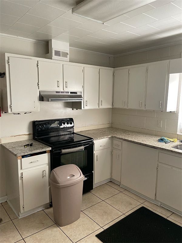 White kitchen cabinets and counters, black range, beige trash can, tiled floor.