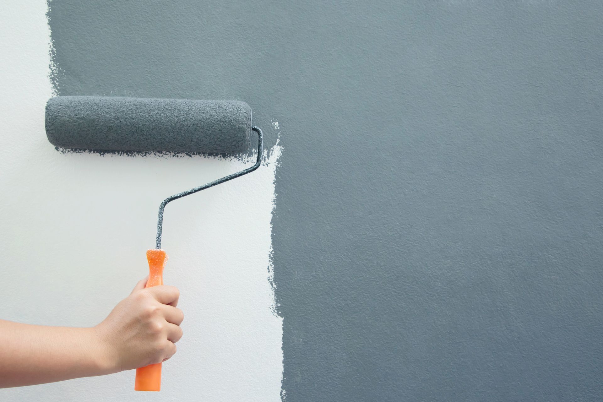 A hand holding a paint roller applies gray paint to a white wall.