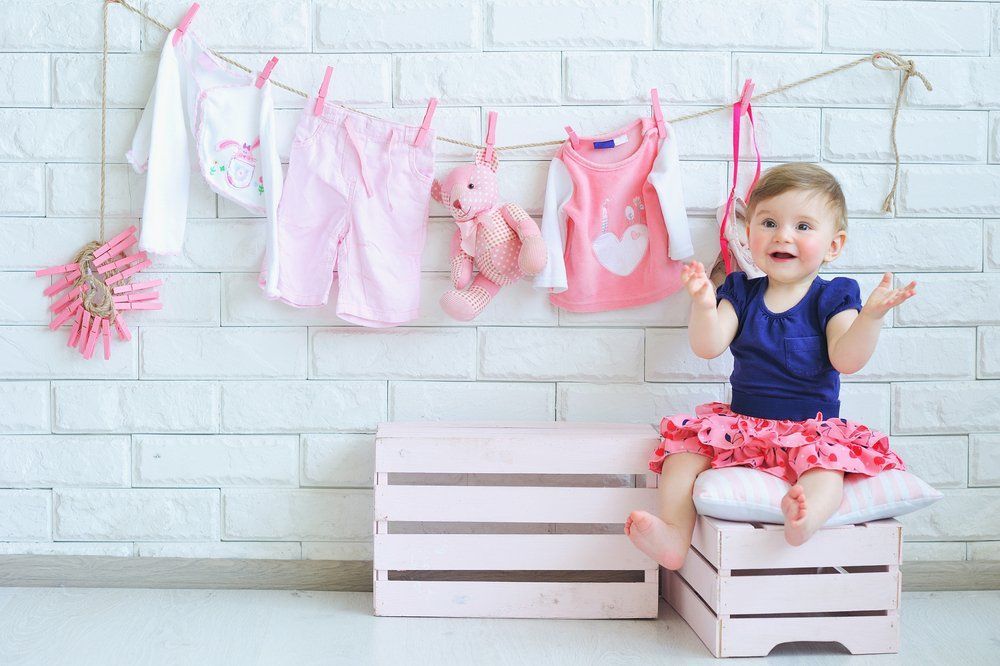 A baby girl is sitting on a wooden box next to clothes hanging on a clothes line.