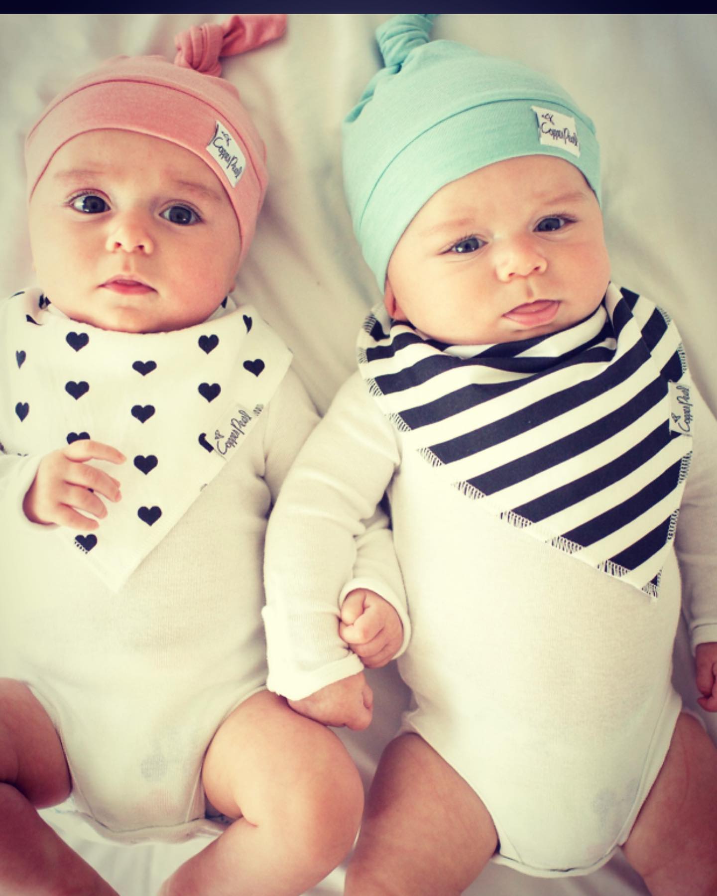 Two babies wearing bibs and hats are laying on a bed