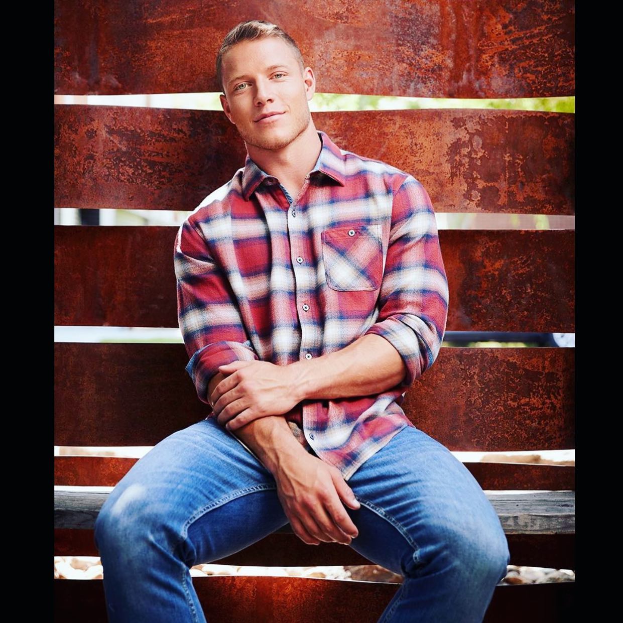 A man in a plaid shirt and jeans is sitting in front of a rusty wall.