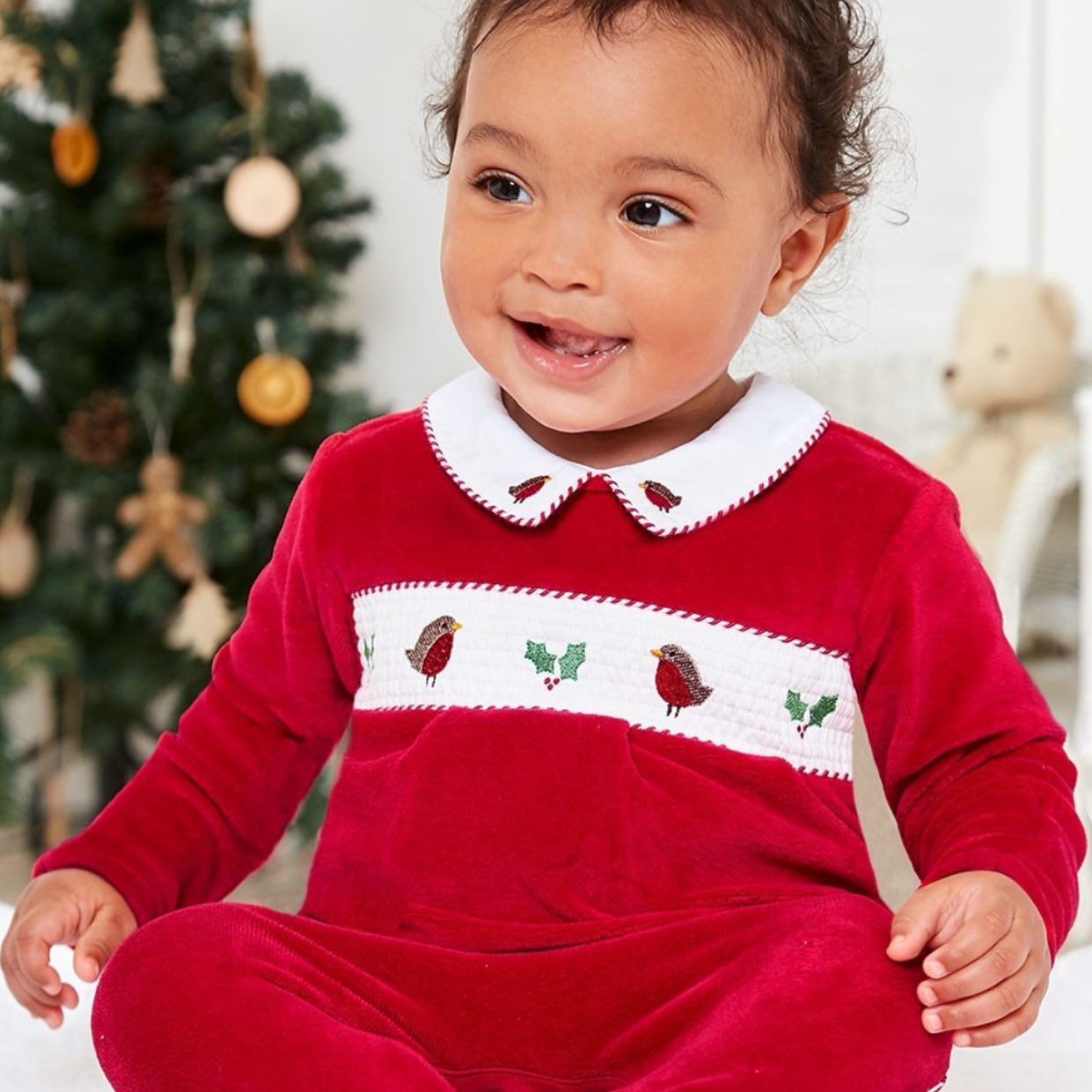A baby in a red dress is sitting in front of a christmas tree
