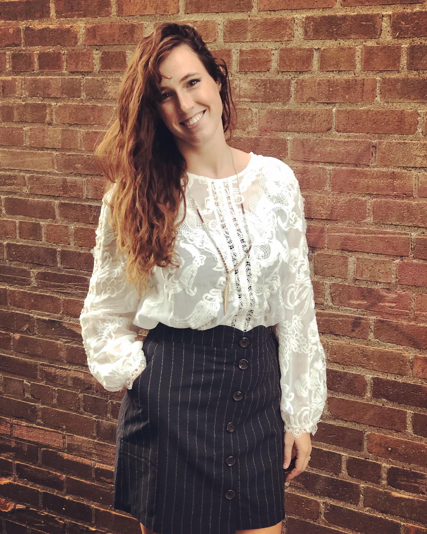 A woman wearing a white blouse and a black skirt is standing in front of a brick wall.