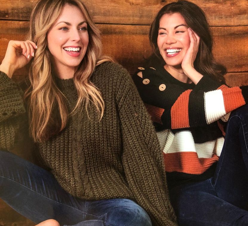 Two women wearing sweaters are sitting next to each other and smiling