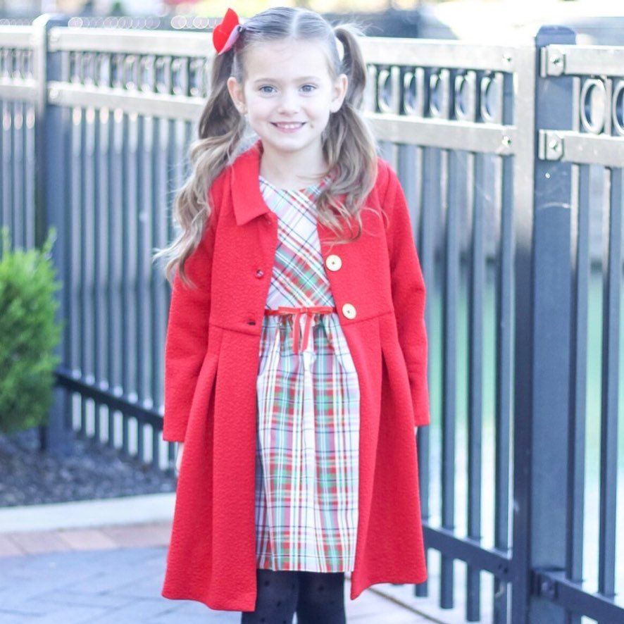 A little girl wearing a red coat and plaid dress is standing in front of a fence.