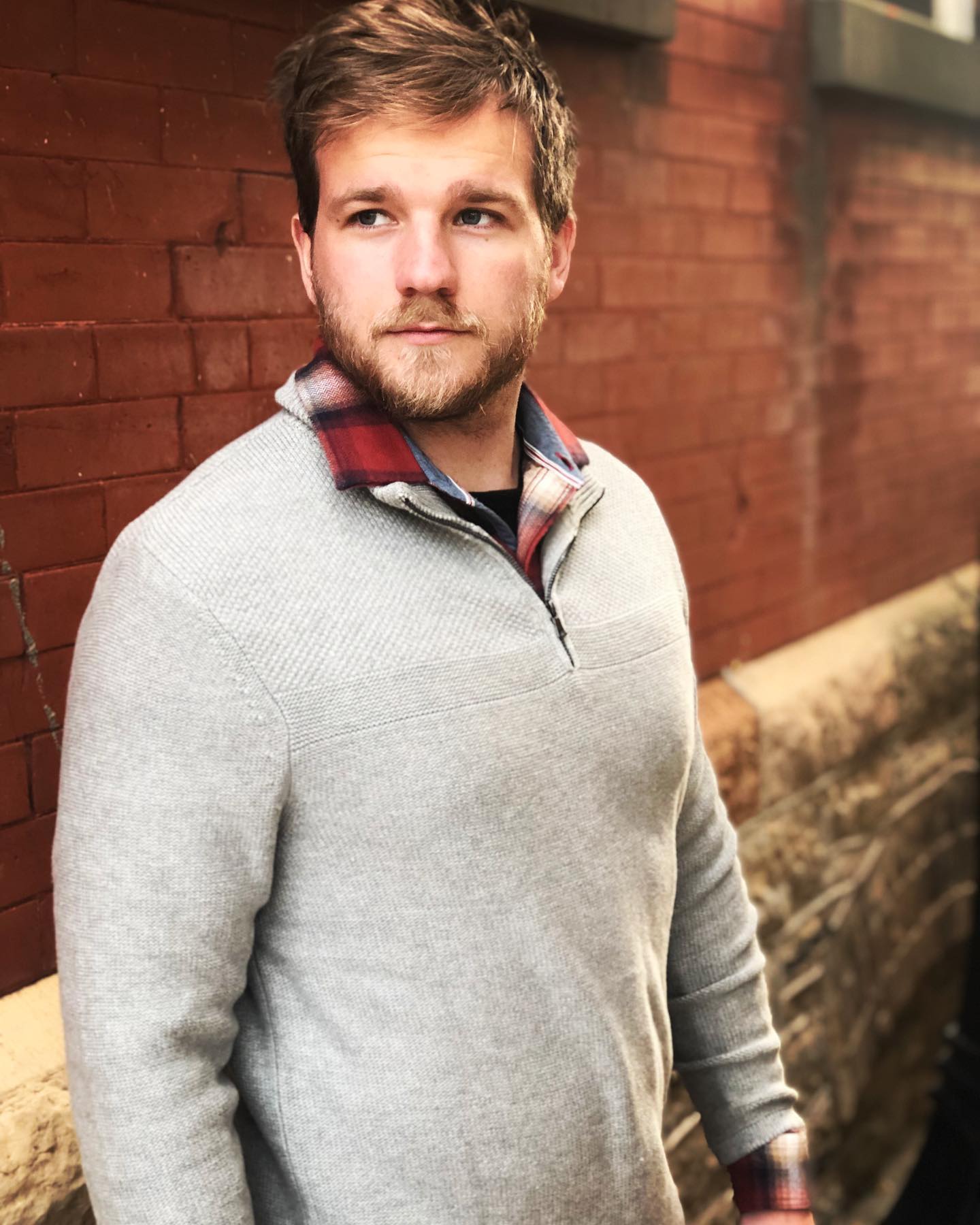 A man wearing a grey sweater and a plaid shirt is standing in front of a brick wall.