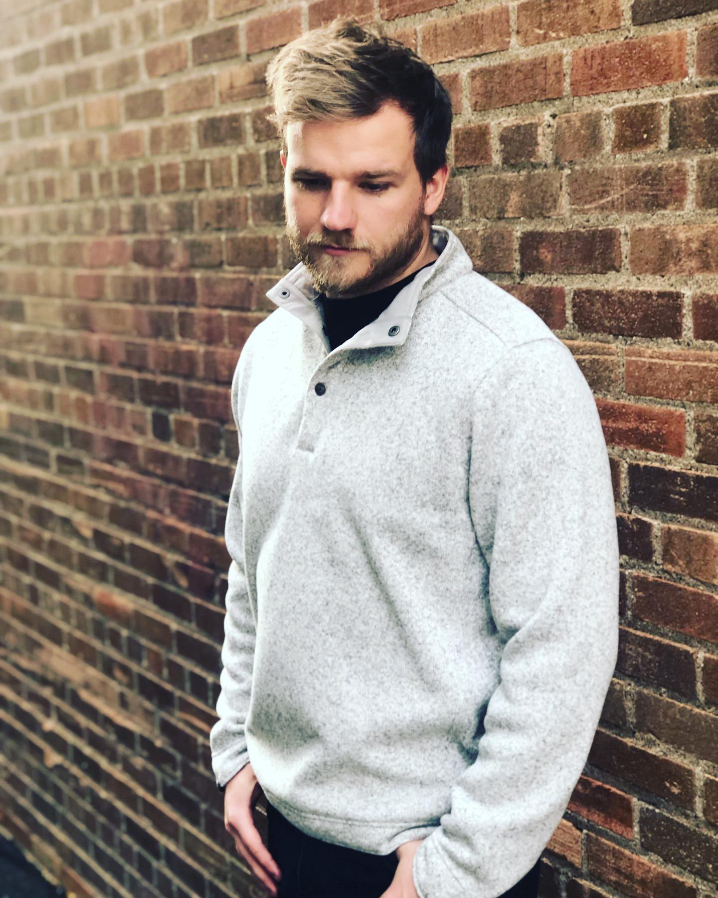 A man in a grey sweater is leaning against a brick wall.