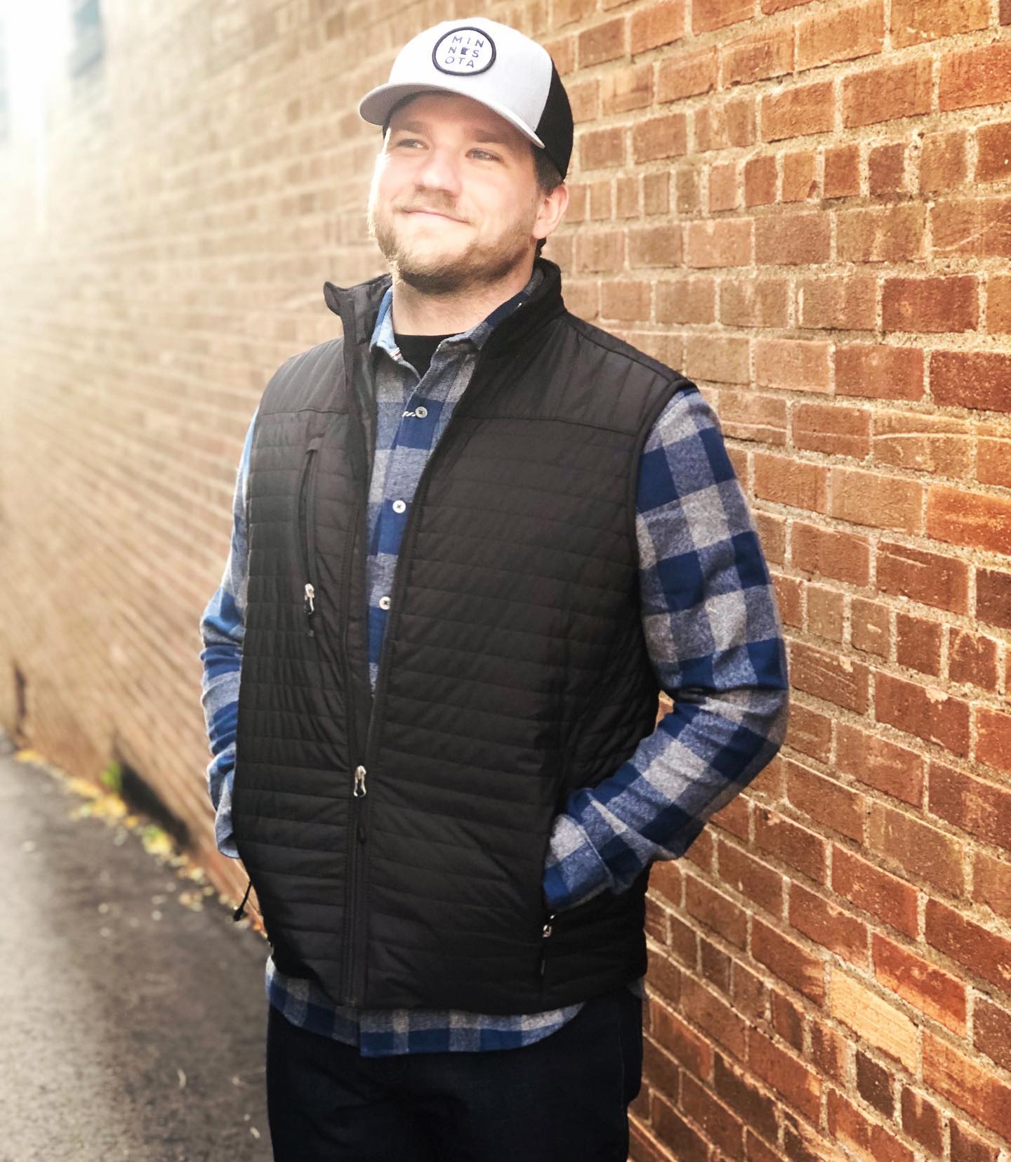A man wearing a black vest and a hat is leaning against a brick wall.