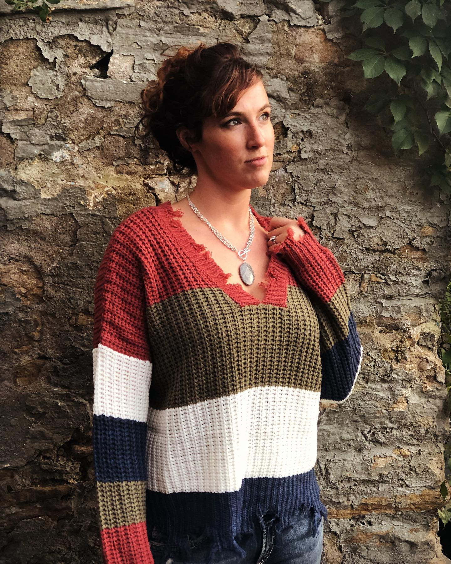 A woman wearing a sweater and a necklace is standing in front of a brick wall.