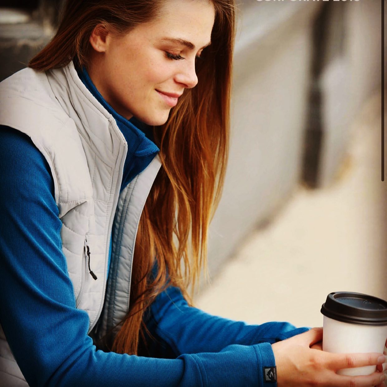 A woman wearing a blue sweater and a grey vest is holding a cup of coffee